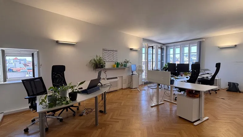 Geräumiges Büro mit Fischgrätparkett, mehreren Arbeitsplätzen und Blick auf die Stadt durch die Fenster.
