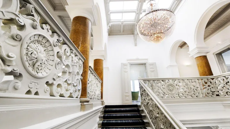 Elegantes Treppenhaus mit kunstvollen Geländern, hohen Decken und einem prächtigen Kronleuchter unter einem Glasdach.