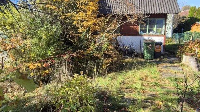 Kleines Haus mit Satteldach, umgeben von herbstlicher Vegetation und Bäumen unter blauem Himmel.