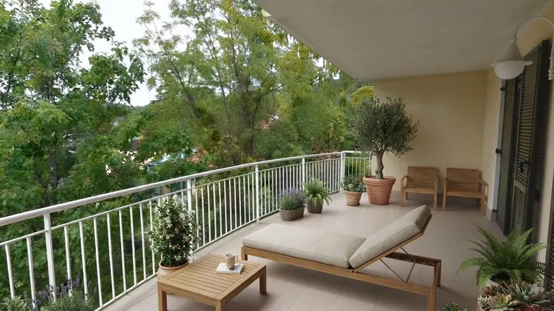 Geräumiger Balkon mit bequemer Sonnenliege, kleinem Tisch und üppiger Bepflanzung, umgeben von grünen Bäumen.