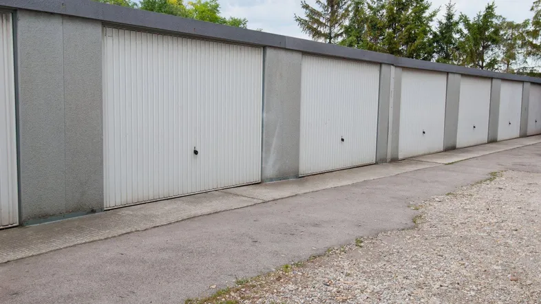 Reihe von Garagen mit weißen Toren und Betonfassade, umgeben von Bäumen und einem Schotterweg.