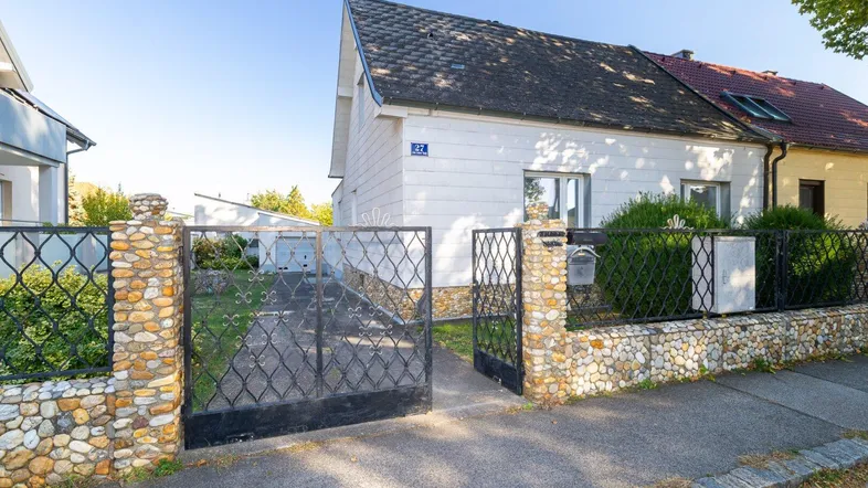 Vorderansicht des Hauses mit schmiedeeisernem Tor und Steinpfeilern, umgeben von Grün.