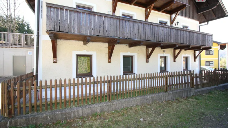 Traditionelles Gebäude mit Holzbalkon und einem kleinen Zaun im Vorgartenbereich, umgeben von Grün.