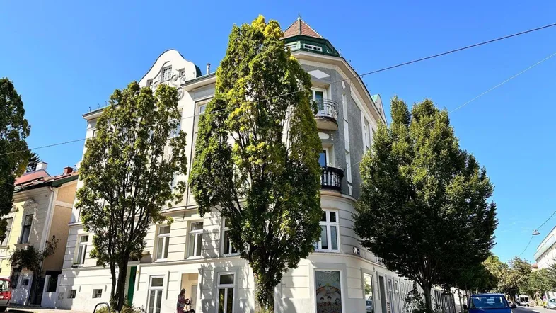 Historisches Gebäude mit klassischer Architektur und hohen Bäumen an einer sonnigen Straße.