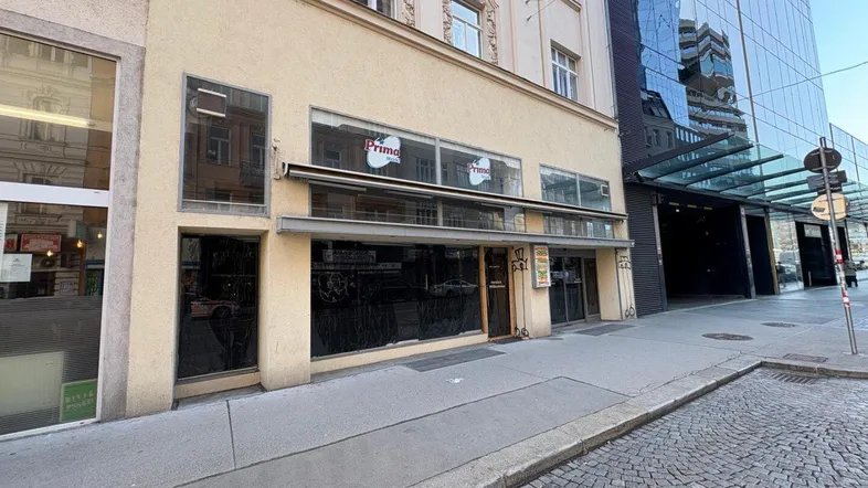Gebäudefassade mit großen Schaufenstern und einem modernen Glasgebäude daneben, an einer städtischen Straße gelegen.