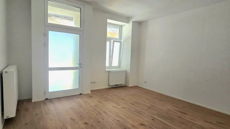 Heller, leerer Raum mit Holzboden, weißen Wänden und Zugang zu einem kleinen Balkon oder einer Loggia.