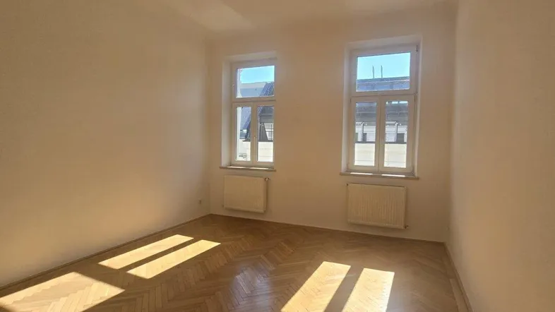 Helles Zimmer mit Fischgrätparkett und zwei Fenstern, durch die Sonnenlicht einfällt.