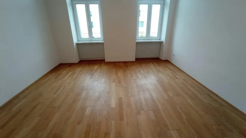 Helles Zimmer mit Holzboden und zwei Fenstern, ideal als Schlaf- oder Wohnbereich.