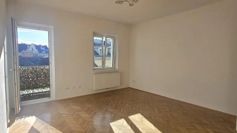 Helles Zimmer mit Fischgrätparkett und Zugang zu einem Balkon, durchflutet von Sonnenlicht.