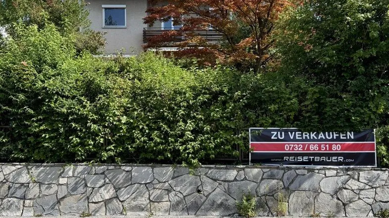 Hausfassade hinter einer Steinmauer und Hecke mit einem 'Zu Verkaufen'-Schild und Herbstbaum.