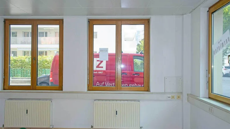 Heller Büroraum mit Laminatboden, großen Holzfenstern und Heizkörpern, der viel Tageslicht hereinlässt.