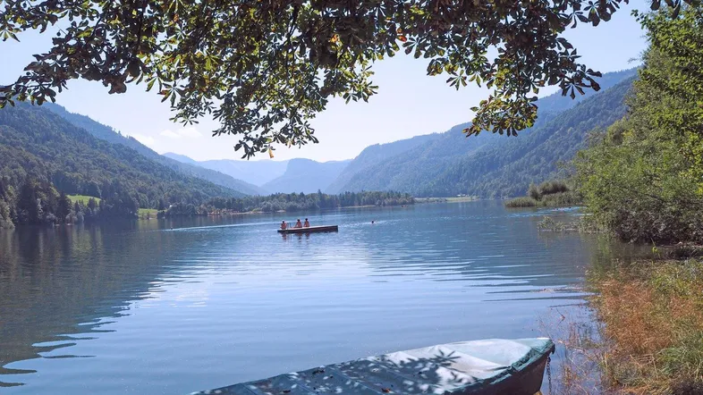 Idyllischer Blick auf einen ruhigen See mit Ruderbooten, umgeben von bewaldeten Bergen unter blauem Himmel.