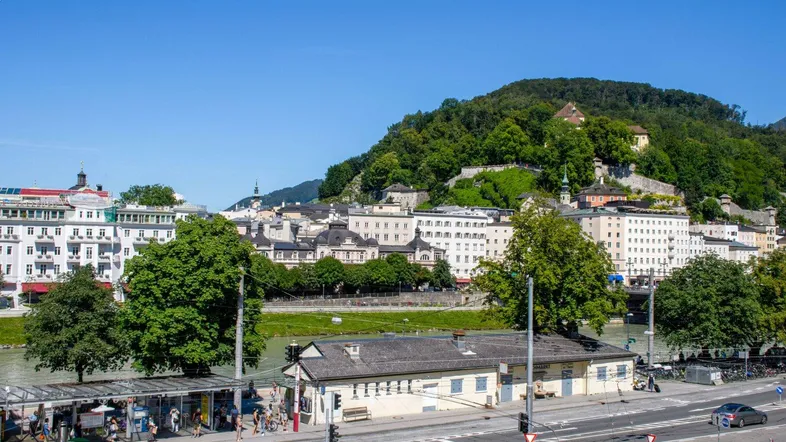 Panoramablick auf die Stadt Salzburg mit Fluss, grünen Hügeln und historischer Architektur unter blauem Himmel.