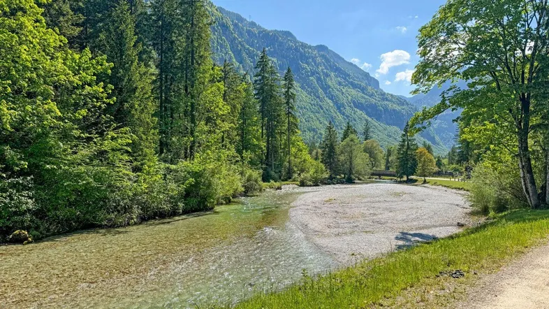 Idyllische Flusslandschaft mit klarem Wasser, umgeben von grünen Wäldern und majestätischen Bergen unter blauem Himmel.