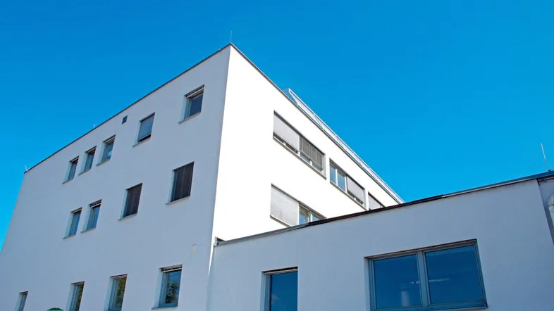Modernes weißes Gebäude mit klaren Linien und Rollläden unter strahlend blauem Himmel.