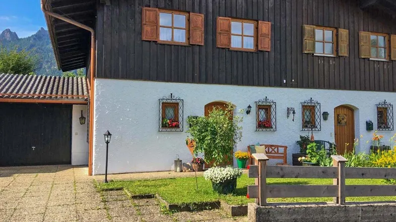 Traditionelles Haus mit Holzelementen und weißer Fassade, umgeben von einem kleinen Vorgarten und Bergblick.