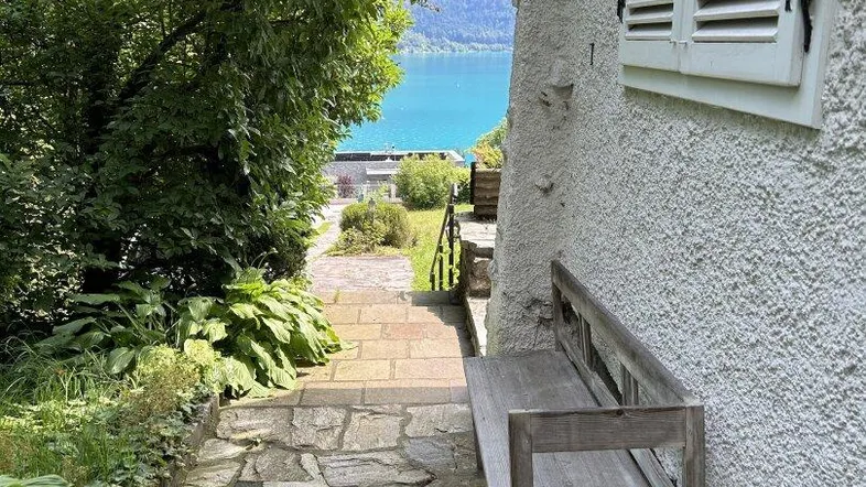 Steinweg führt zu einem Seeufer mit einer Holzbank neben dem Haus, umgeben von üppigem Grün.