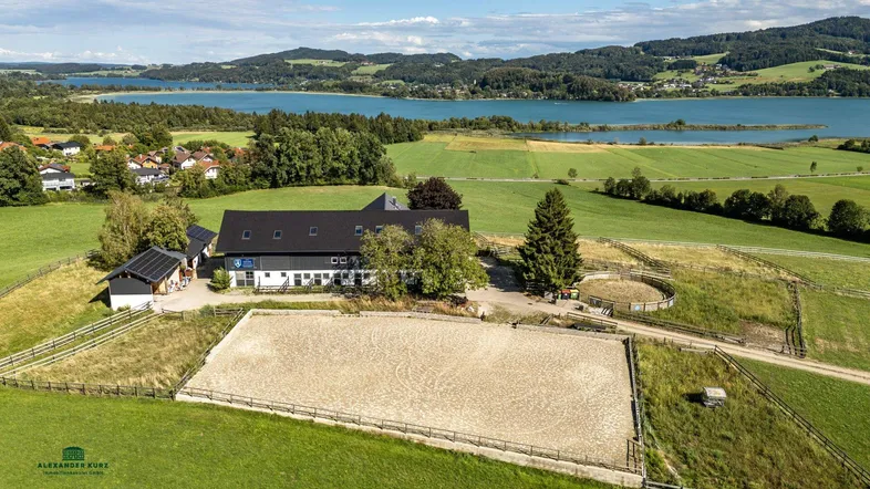 Luftaufnahme eines Anwesens mit Reitplatz, Stallungen und malerischem Seeblick in ländlicher Umgebung.