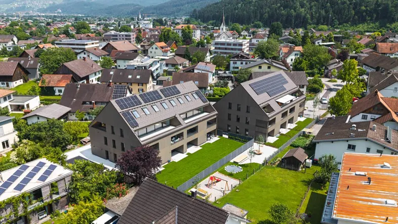 Luftaufnahme von modernen Neubauten mit Photovoltaikanlagen, Gärten und Spielplatz in einer grünen Wohngegend mit Bergblick.