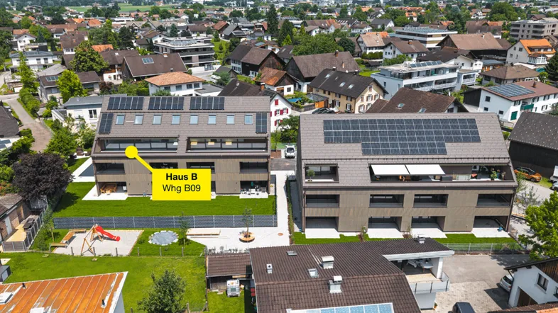 Luftaufnahme von modernen Neubauten mit Photovoltaikanlagen, umgeben von grünen Gärten und einem Spielplatz, mit Bergblick im Hintergrund.