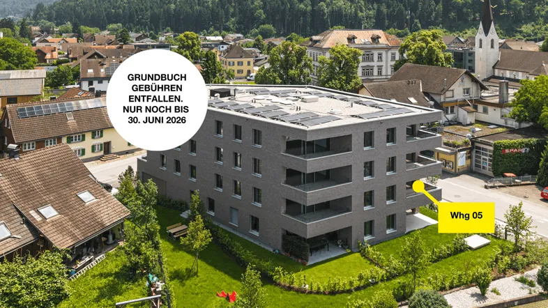 Modernes Mehrfamilienhaus mit Flachdach und Solaranlage, umgeben von Grünflächen und einer ländlichen Umgebung mit Bergblick.