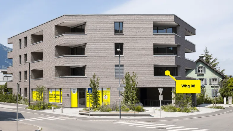 Modernes Mehrfamilienhaus mit grauer Fassade, Balkonen und überdachten Parkplätzen an einer Straßenecke mit Bergblick.