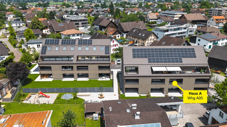 Luftaufnahme von modernen Mehrfamilienhäusern mit Solaranlagen auf den Dächern, umgeben von viel Grün und einem Spielplatz, mit Bergblick.