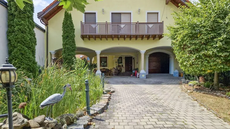 Gelbes Haus mit gepflasterter Auffahrt, kleinem Teich und einem Balkon im Obergeschoss.