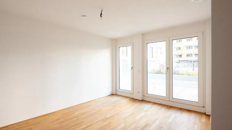 Heller, leerer Raum mit Holzboden und großen Fenstern, die viel Tageslicht hereinlassen.