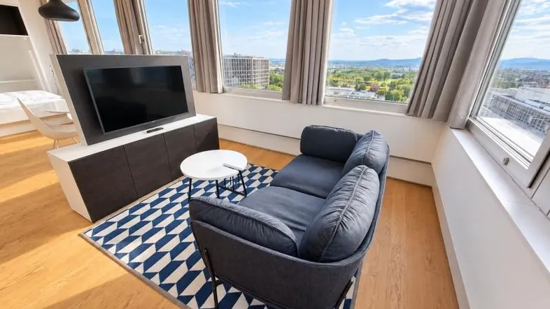 Helles Wohnzimmer mit bequemer Couch, Fernseher und weitem Blick über die Stadt und die grüne Landschaft.