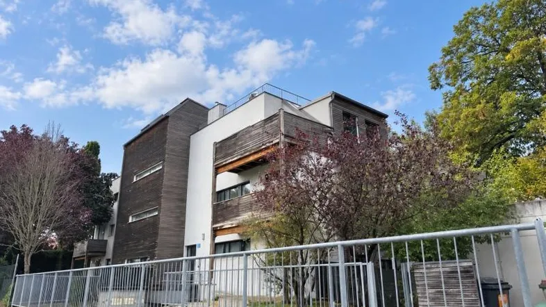 Modernes Gebäude mit Holz- und Weißfassade, umgeben von Bäumen und einem Zaun.