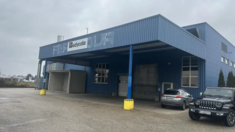 Industrielles Gebäude mit blauer Wellblechfassade und überdachtem Eingangsbereich, davor geparkte Fahrzeuge auf dem Vorplatz.