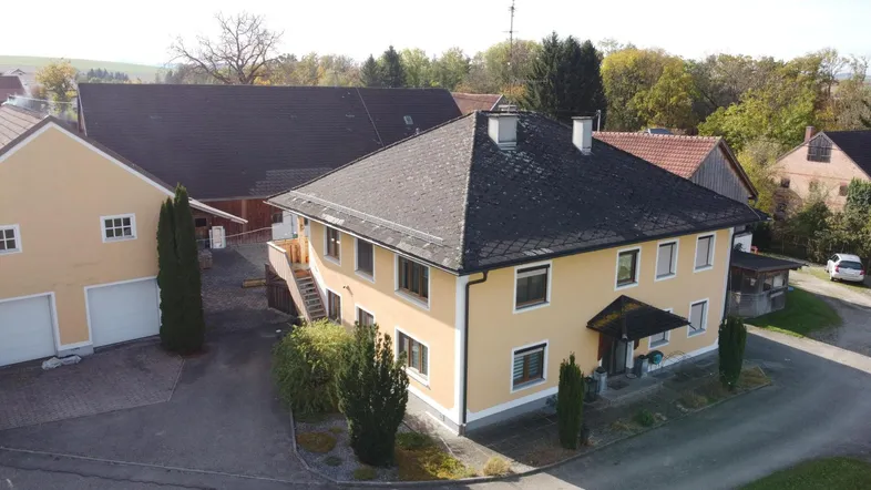 Luftaufnahme eines gelben Wohnhauses mit dunklem Dach und umliegender ländlicher Bebauung.
