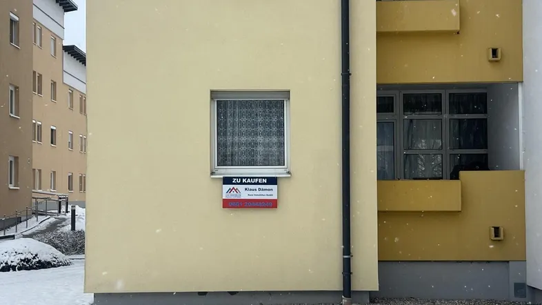 Gelbe Fassade eines Mehrfamilienhauses im Winter mit einem 'Zu Kaufen'-Schild am Fenster.