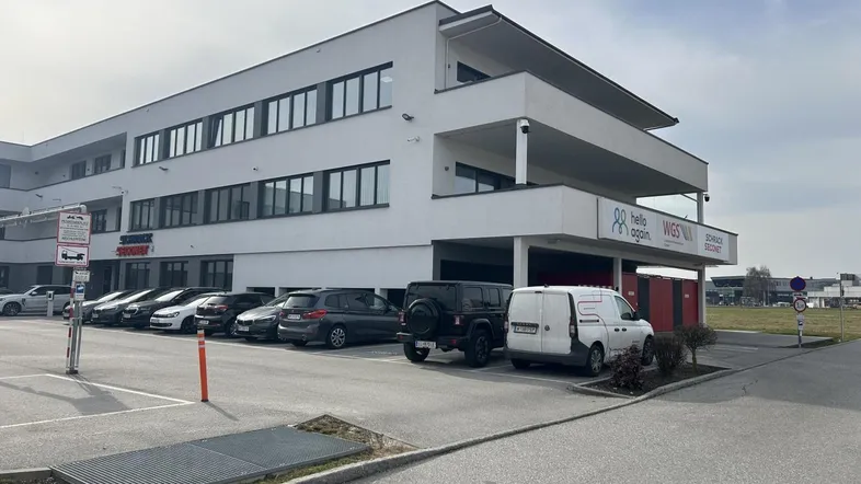 Modernes Geschäftsgebäude mit weißer Fassade und großem Parkplatz unter bewölktem Himmel.