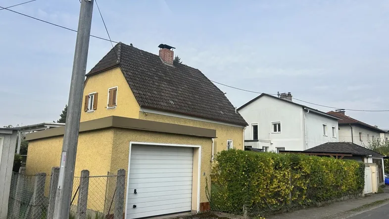 Gelbes Einfamilienhaus mit Satteldach und integrierter Garage, umgeben von einer grünen Hecke.