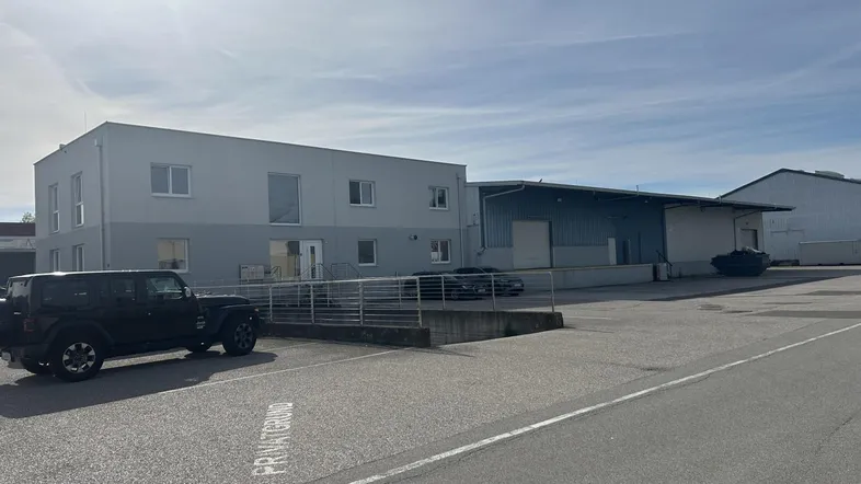 Außenansicht eines Gewerbegebäudes mit Bürotrakt und Lagerhalle, umgeben von einem großen Parkplatz.
