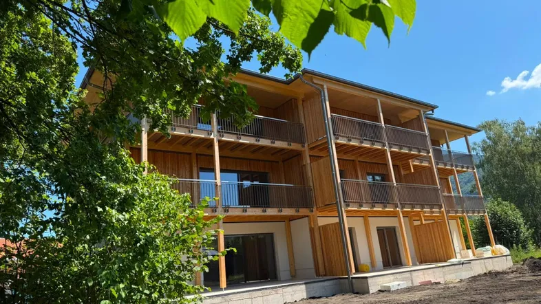 Neubau mit Holzfassade und mehreren Balkonen, umgeben von viel Grün unter blauem Himmel.
