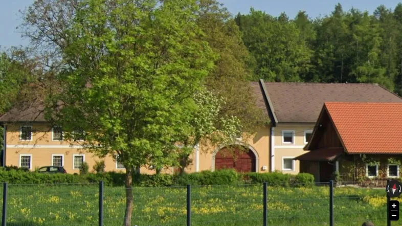 Traditionelles Gebäude mit gelber Fassade und braunem Dach, umgeben von viel Grün und Bäumen.