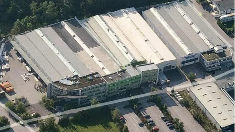 Luftaufnahme eines großen Gewerbekomplexes mit mehreren Hallen, Bürogebäuden und Parkplätzen, umgeben von Bäumen.