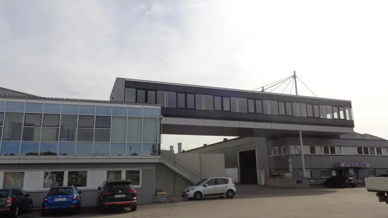 Industriekomplex mit mehreren Gebäuden und einer erhöhten Verbindungsbrücke, umgeben von einem Parkplatz.