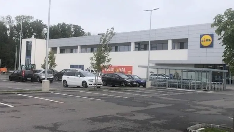 Moderne Außenansicht des Lidl-Gebäudes mit großem Parkplatz und Bäumen im Vordergrund.