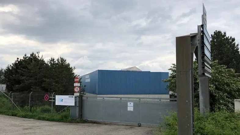 Außenansicht eines Industriegebäudes mit blauer Fassade, umzäuntem Gelände und einem Eingangstor unter bewölktem Himmel.