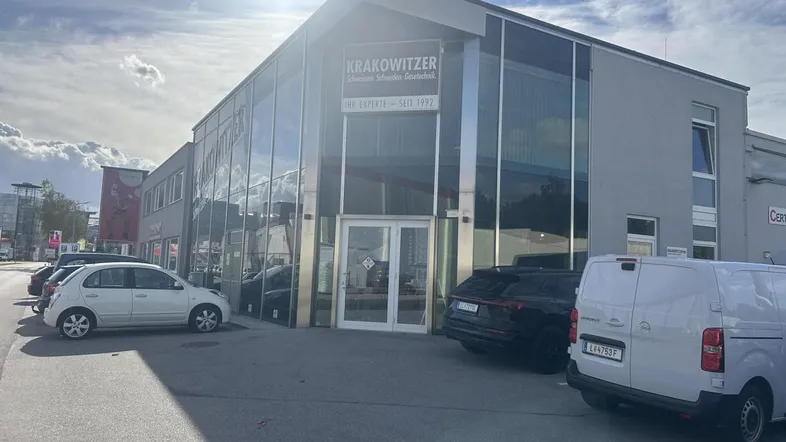Modernes Geschäftsgebäude mit großer Glasfassade und Parkplätzen davor unter hellem Himmel.