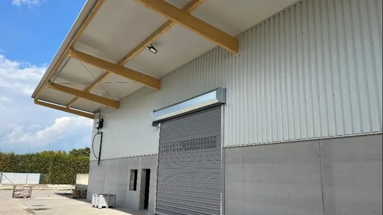 Außenansicht eines modernen Industriegebäudes mit Rolltor, Wellblechfassade und sichtbaren Holzträgern unter dem Dachüberstand.