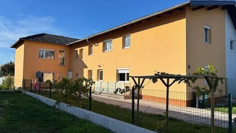 Außenansicht eines Gebäudes mit gelber Fassade, schwarzem Dach und einem kleinen Gartenbereich mit Pergola.