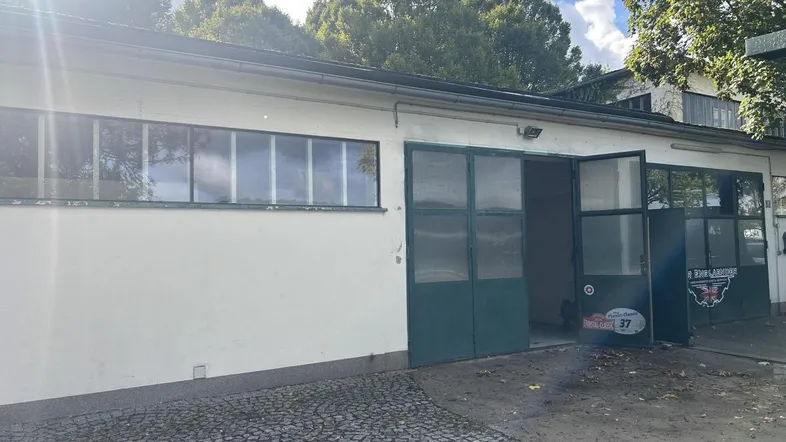 Außenansicht einer Werkstatt mit großen Toren und Fenstern, umgeben von Bäumen und einem gepflasterten Vorplatz.