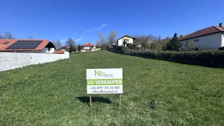 Grünes Grundstück mit 'Zu verkaufen'-Schild und umliegenden Häusern unter blauem Himmel.