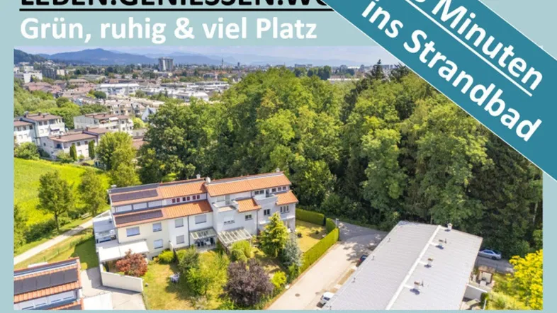 Luftaufnahme eines Mehrfamilienhauses mit Garten, umgeben von viel Grün und Bergen im Hintergrund.