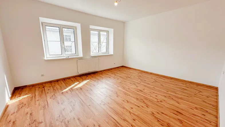 Helles, leeres Zimmer mit Holzfußboden und zwei Fenstern, bereit zur individuellen Gestaltung.